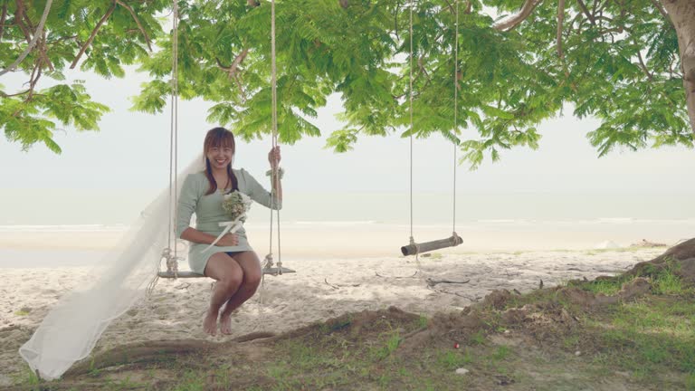 Scene of Asian bride holding the flower on her hand with smiling face sitting on the swing near the beach