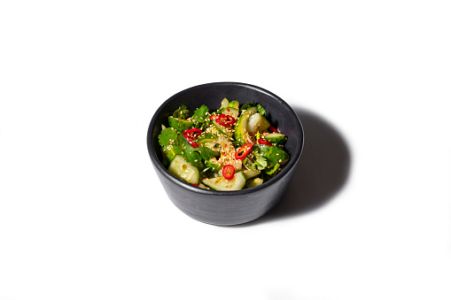 Asian cucumber salad with sesame seeds, chilli rings and sweet and sour sauce. The salad lies in a black, ceramic bowl. White background
