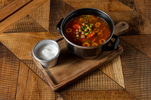 Meat hodgepodge with vegetables. Olives, capers, tomatoes, veal, carrots, salami. Soup in a black enamel pot. Nearby is a metal, small bowl with sour cream. Soup and sour cream stand on a wooden board. The board stands on a light, wooden table with a pattern.