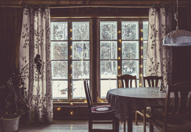 Wooden grid pattern window in cottage home. Dark, light coming from windows, snowy trees on background. Wood table with chairs indoors.