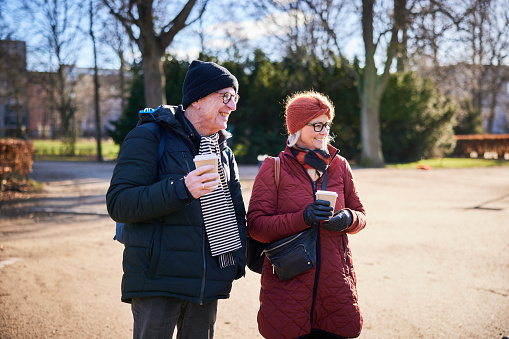 Smiling senior couple drinking takeaway coffee while walking together in a city park on a sunny day in winter