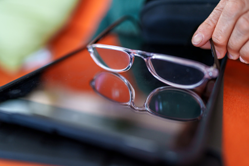 The elderly woman is cleaning her reading glasses to use her digital tablet.