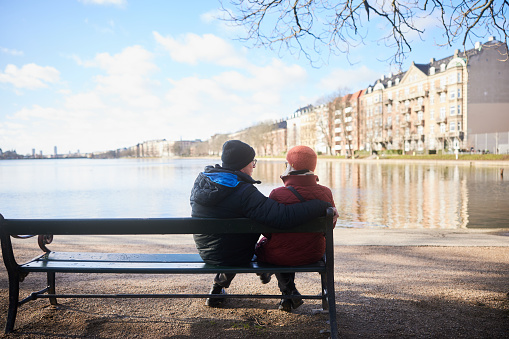Rear view of a senior couple sitting side by side together on a bench in a park by a river in winter