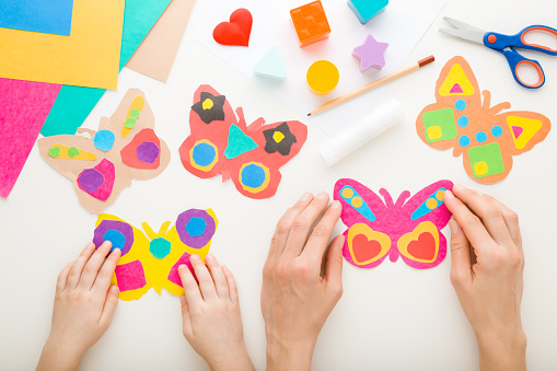 Young adult mother and child hands holding colorful paper butterfly shapes on white table background. Point of view shot. Closeup. Playing and spending time together. Top down view.