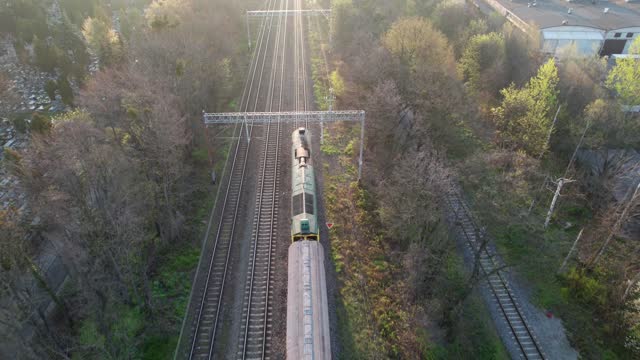 A drone-shot captures a freight train passing through the city in the morning, crossing a viaduct and green landscapes. The locomotive pulls empty wagons to be filled with raw materials.