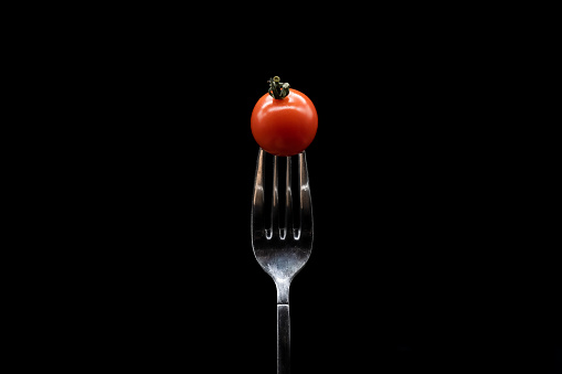Isolated cherry tomato prickled by a silver fork on black background. Minimalist picture of mediterranean diet.