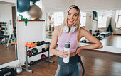 Cheerful blonde caucasian young girl in sportswear holds bottle of water looks at camera toothy smiles stands at fitness class. Healthy lifestyle, sporty women. American woman at break of exercise.