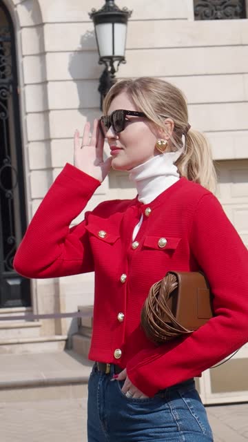 Outdoor portrait of young elegant fashionable woman puts on sunglasses, wears colorful red blazer, leather bag, turtleneck stands in European city street. Clothes Trends of spring and summer
