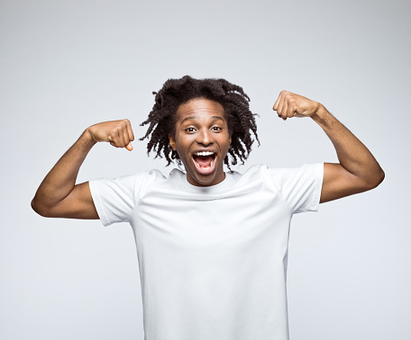 Excited afro american young man wearing white t-shirt, laughing at camera and flexing his muscles. Studio shot on grey background.