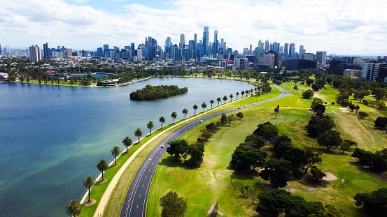 Albert Park Lake, Melbourne City, Victoria, Australia. Aerial drone view of the lake with a row of palm trees, the road, the open-wheel single-seater racing car grand prix racing track, walking track and bike riders. Sky-rise buildings and the melbourne skyline in the background.