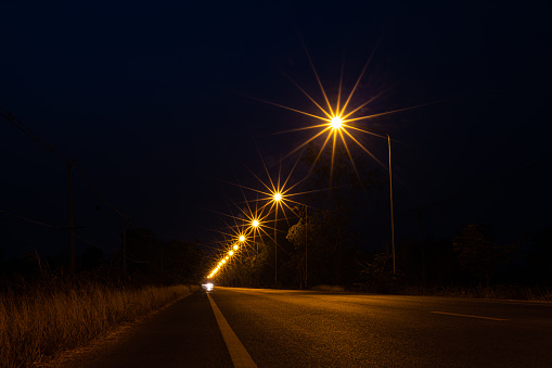 A view of orange starbursts from a lamppost on the side of a street illuminating the tarmac and dry grass at night with a dark sky in the background with headlights lit in the distant Thai countryside.