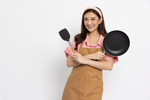 Young Asian woman housewife wearing kitchen apron cooking and holding pan and flipper isolated on white background