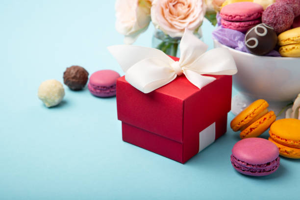 Sweet cookies macaroons, red gift box, rose flowers and chocolate candies on blue background. Spring presents concept for Mothers day, womans day, birthday, empty space for text or message