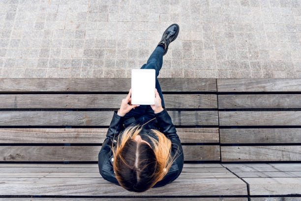 istock Mockup image of woman holding and using white tablet pc with blank white desktop screen while sitting on the wooden bench.Top view.Copy space. 1485118683