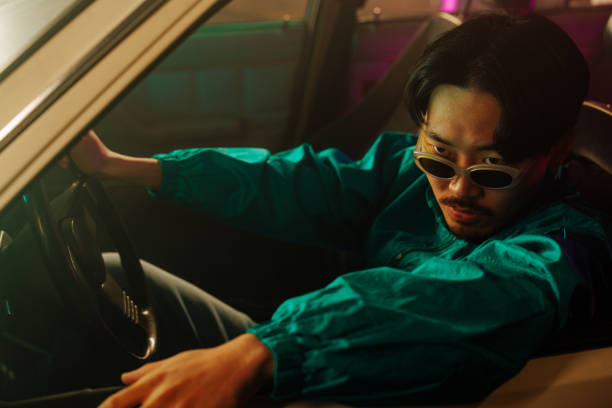 A retro stylish Asian man is sitting in the car and looking at the camera through the window.