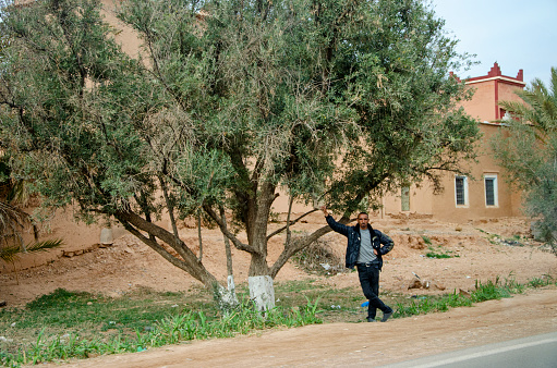 Morocco, road that leads from Taroudant to Ouarzazate. 03/17/2023. Man leaning against a tree in the shade waiting for a ride. Rural landscape of Morocco
