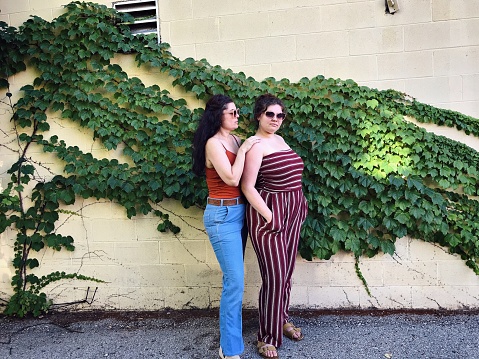 Beautiful plus size body positive and confident Gen Z girl and her mother playing around and posing and shopping in downtown Iron Mountain, Michigan in the business district. They are happy and carefree posing among the buildings and ivy growing up around the buildings on a city outing day, spending quality time together.