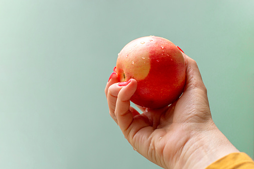 Female hand holding apple against pastel green background with copy space. The apple is washed and drops of water are visible.