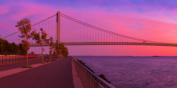 High resolution stitched panorama of the Verrazano-Narrows Bridge at Red Orange Yellow Blue Majestic Sunset. The bridge connects boroughs of Brooklyn and Staten Island in New York City. The bridge was built in 1964 and is the largest suspension bridge in the USA. Historic Fort Wadsworth is next to the foot of the bridge. The photo was taken from the Shore promenade in Bay Ridge Brooklyn. Canon EOS 6D full frame censor camera. Canon EF 85mm F/1.8 Prime lens. 2:1 Image Aspect Ratio. This image was downsized to 50MP.