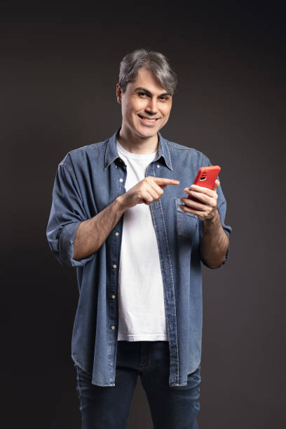 retrato de hombre brasileño, con camisa abotonada y jeans, usando teléfono inteligente y mirando hacia adelante, posición 3/4, cabello gris - foto en color - brasil - handsome man flash fotografías e imágenes de stock