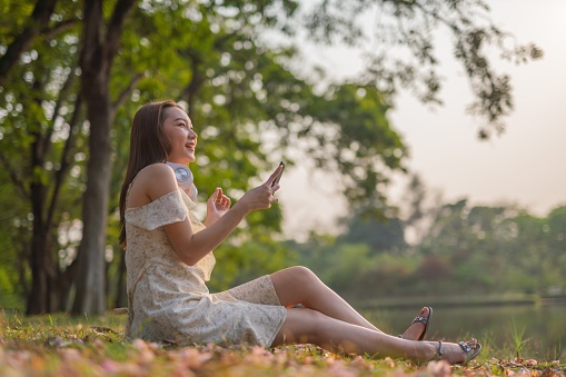 Landscape portrait photo shot of a young asian woman sitting on the ground in a beautiful park using her phone and listening to music