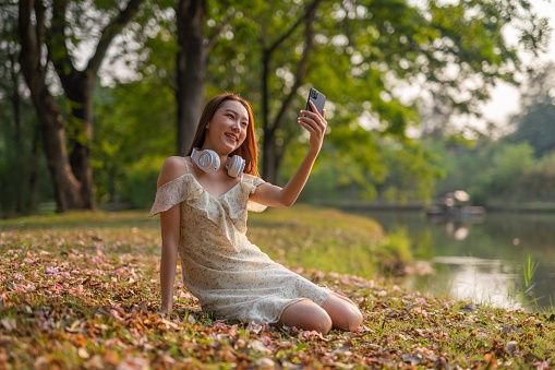Landscape portrait photo shot of a young asian woman sitting on the ground in a beautiful park posing for selfie and listening to music