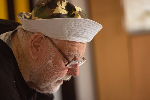 Shot of a senior man with dementia, wearing glasses and hat, looking down.
