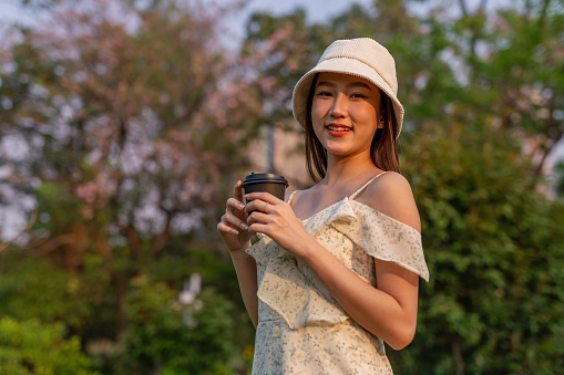 Half body shot of a young beautiful asian woman holding a cup of coffee wandering in a park outdoors during a sunset moment