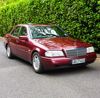Cape Town, South Africa - February 5, 2023: Side view of a well-preserved late-1990s Mercedes-Benz 230K Sport in deep metallic maroon, against a lush hibiscus hedge, parked in a street in a wealthy suburb of Cape Town, South Africa.
