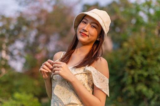 Half body shot of a young beautiful asian woman with a hat wandering in a park outdoors during a sunset moment