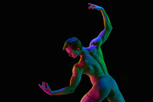 Contemporary dance. Portrait with young, handsome, sporty and athletic ballet dancer posing over dark studio background with neon light. Back view. Grace, art, professional sport concept