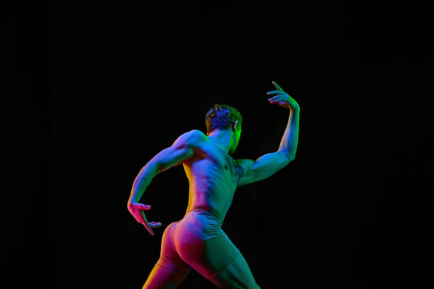 Power, freedom. Young flexible athletic man, handsome dancer posing with hands up on dark studio background with neon light. Modern ballet style. Grace, art, professional sport concept