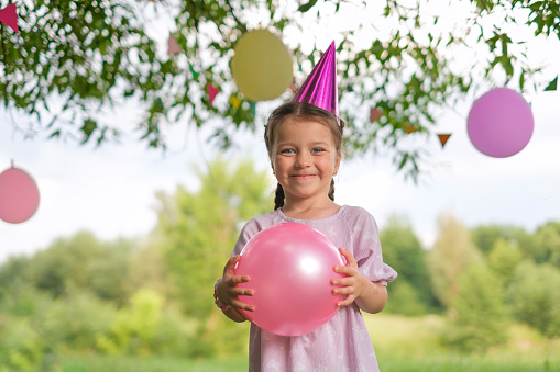 A girl in a festive hat stands with a ball smiling on her birthday. Balloons and decorations for the holiday hang on the trees