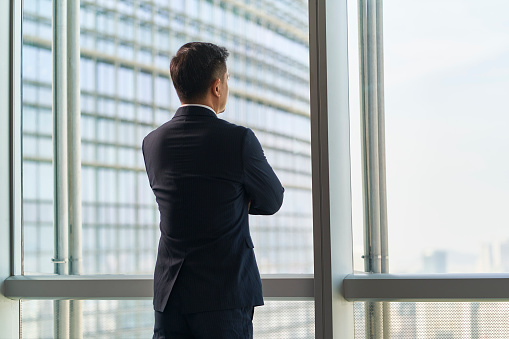rear view of asian business man standing by the window looking at city view arms crossed