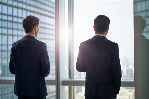 rear view of two asian business men standing by the window in office looking at the city talking chatting in the morning sun