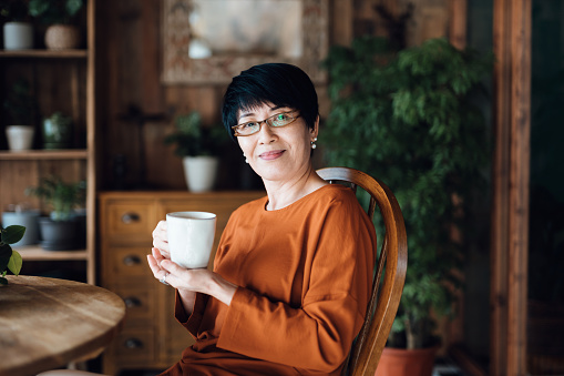 Portrait of senior Asian woman enjoying a cup of coffee, sitting at dining table against green plants, relaxing at home. Elderly and retirement lifestyle