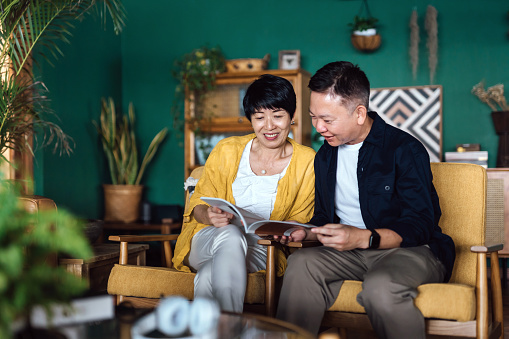 Happy senior Asian couple sitting on armchair in the living room, enjoying time together, relaxing and reading a travel magazine. Elderly and retirement lifestyle