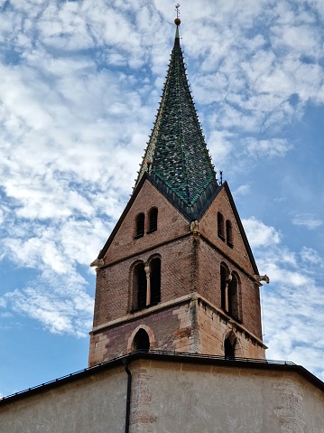 Pointed church tower in Trentino with polychrome tile roof against blue sky with light white clouds
