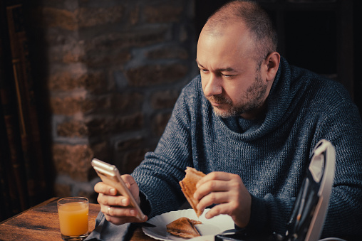 A man eats a sandwich and looks at the phone at home. Ordinary middle-aged man. Eating with a phone in hand.