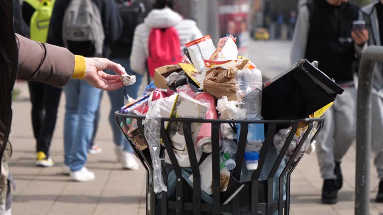 Hand throwing a piece of paper into the overflowing garbage bin in the city with blurred and unrecognizable people walking in the background