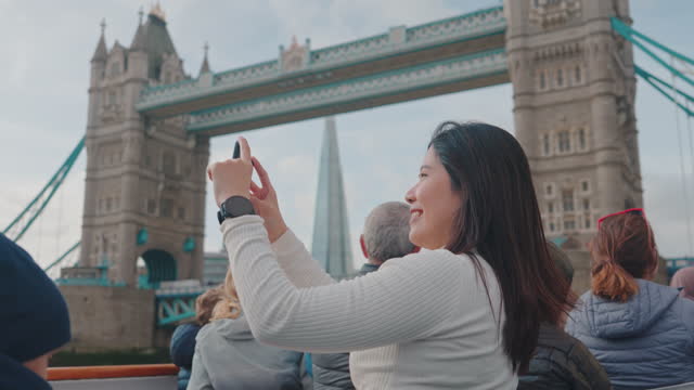 Asian woman tourist using mobile phone and taking photo while on River Thames boat tours with Tower Bridge view in London, United Kingdom