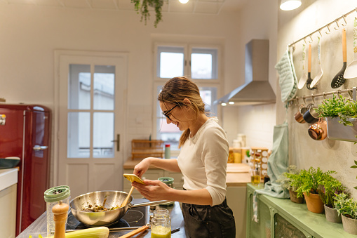 Photo of a young woman busy in her home kitchen, preparing dinner for herself and connecting with her family members over a video call while cooking.