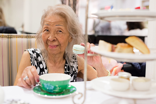 Elderly Filipino woman having tea with desserts and looking at the camera while holding a macaroon