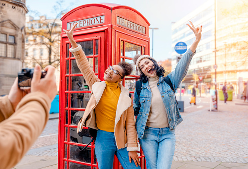 two friend, girlfriend and women using a mobile phone, camera and taking selfie against a red phonebox in the city of England.Travel Lifestyle concept