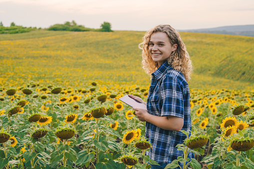 Woman farmer in workwear standing in front of camera and using touchpad standing in sunflower field. Smiling woman outdoor portrait. Beauty portrait.