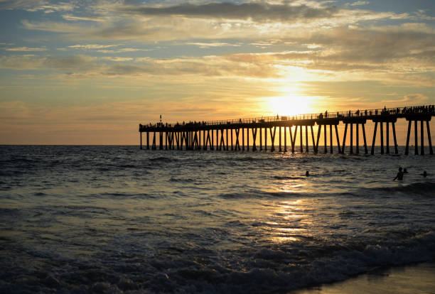 A sunset at Hermosa Beach, California during the summer. The golden sun shines its light on the water's surface as it sets. Swimmers play in the water's waves and splash around. Tourists and locals alike are silhouetted walking on the pier. The Hermosa Beach Pier is silhouetted by the sun. The sunset is comprised of hues of peach, baby blue, cyan, and blue. The puffy white clouds hang low over the ocean.