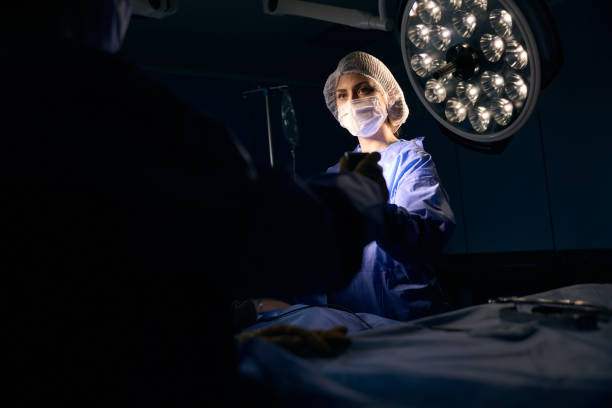 Female in a protective mask stands at the operating table Female in a protective mask stands at the operating table, she passes the instrument to her colleague surgical light stock pictures, royalty-free photos & images