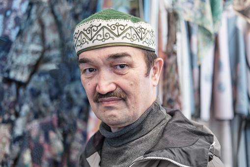 Close-up portrait of senior Asian man looking at camera wearing skullcap made of soft felt and decorated with national ornaments. Kyrgyz headdress topu.