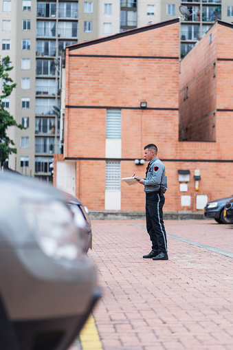 Average age Latino man in his 30s dressed in a security guard uniform is inside the building he works for taking bills for vehicles parked in the parking lot