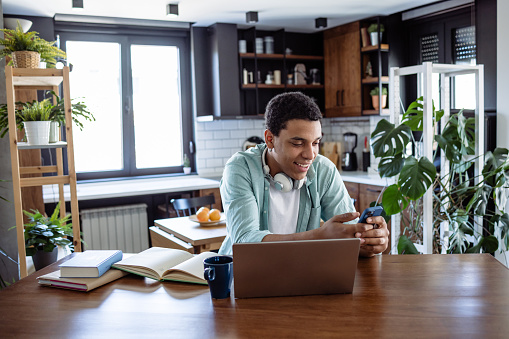 Portrait of a young African American man using a mobile phone while taking a break from working. He is sitting at the table and wearing smart casual clothes. Positive emotion.